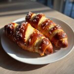 Two freshly baked pretzel dogs, shiny and golden brown, sprinkled with coarse salt, resting on a white plate.