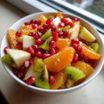 Close-up of a vibrant winter fruit salad featuring orange segments, kiwi, apple chunks, and bright red pomegranate seeds.