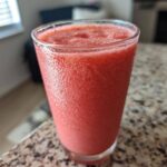Close-up of a tall glass filled with a vibrant, frosty watermelon smoothie sitting on a granite countertop.