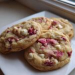 Four freshly baked white chocolate raspberry cookies piled on a white square plate near a window.