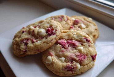 Four freshly baked white chocolate raspberry cookies piled on a white square plate near a window.