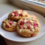 Four freshly baked white chocolate raspberry cookies piled on a white plate near a window.