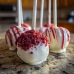A close-up of white chocolate coated red velvet cake pops, one topped with red crumbs, sitting on a granite counter.