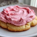 A close-up of a thick, soft Copycat Crumbl Sugar Cookie topped with generous swirls of bright pink frosting.