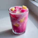 A tall glass filled with a bright pink, icy Copycat Starbucks Dragonfruit Refresher, topped with chunks of dragon fruit and pineapple.