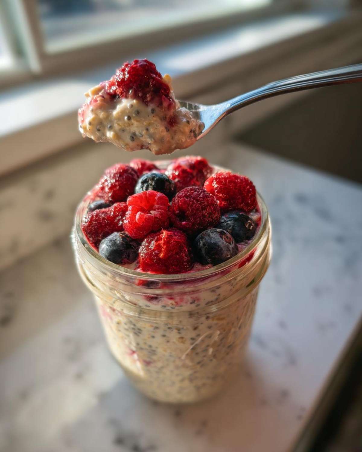 A spoonful of Easy Berry Chia Overnight Oats topped with fresh raspberries and blueberries being lifted from a jar.
