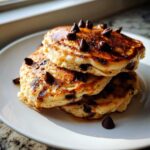 A stack of three fluffy Gluten-Free Chocolate Chip Banana Pancakes drizzled with syrup and topped with chocolate chips.
