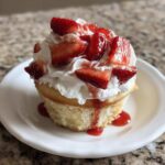 A single Gluten-Free Strawberry Shortcake Cupcake topped with whipped cream and fresh strawberries on a white plate.