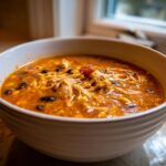 A close-up of a white bowl filled with rich, orange Instant Pot Creamy Chicken Taco Soup featuring shredded chicken, black beans, and corn.