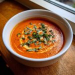 A close-up of a white bowl filled with vibrant orange Instant Pot Creamy Tomato Bisque, swirled with cream and topped with fresh basil.
