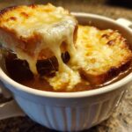 A spoonful of bread topped with melted, gooey cheese is lifted from a bowl of rich Instant Pot French Onion Soup.