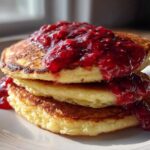 Stack of three golden Keto Coconut Flour Pancakes topped generously with vibrant, chunky raspberry sauce.