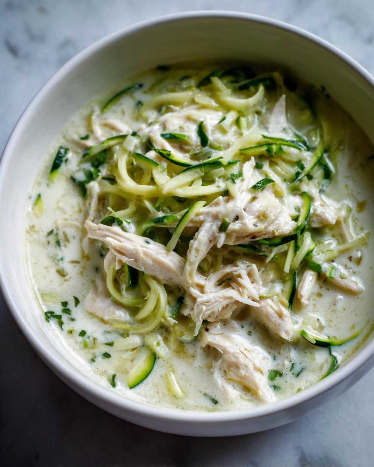 A close-up view of a bowl filled with Keto Creamy Chicken Zoodle Soup, featuring shredded chicken and zucchini noodles in a rich, white broth.