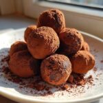 A stack of rich, dark No-Bake Chocolate Orange Truffles heavily dusted with cocoa powder on a white plate.