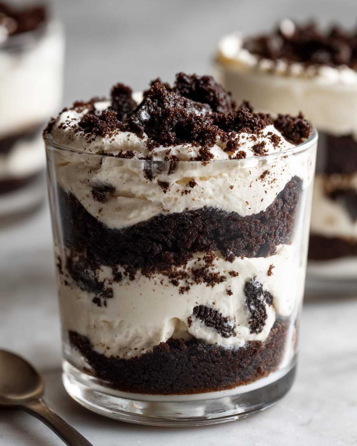 A close-up of a layered No-Bake Cookies and Cream Pie Cups dessert in a glass, featuring dark chocolate cookie crumbs and white cream filling.