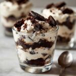 A close-up of a glass filled with layered No-Bake Cookies and Cream Pie Cups, topped with whipped cream and cookie pieces.