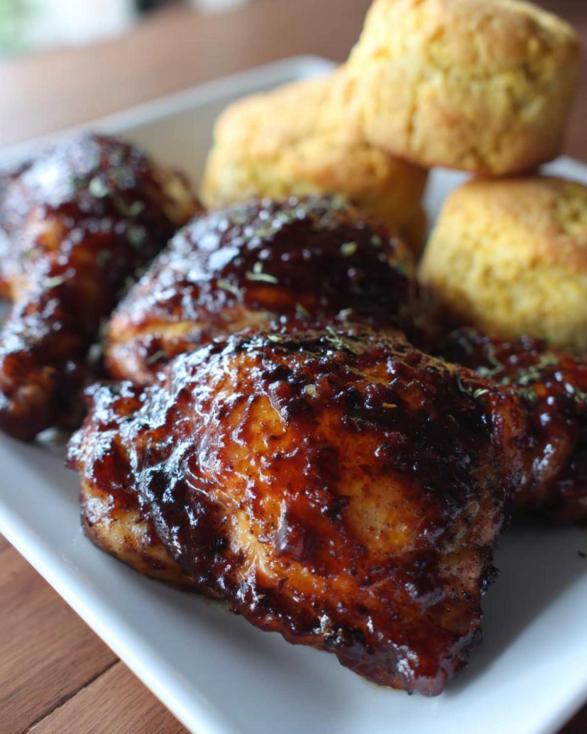 Close-up of glazed BBQ Chicken Thighs served alongside golden Cornbread Biscuits on a white platter.