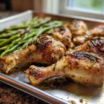 Close-up of golden brown Sheet Pan Lemon Garlic Butter Chicken Legs with herbs on a baking sheet next to roasted asparagus.