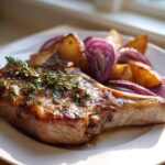 A perfectly cooked Sheet Pan Maple Dijon Pork Chop glazed and topped with fresh thyme, served with roasted red onions and potatoes.