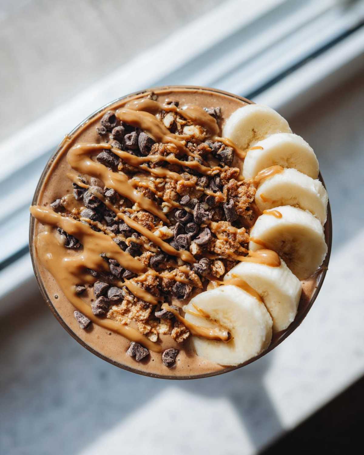 Overhead view of a rich Vegan Chocolate Peanut Butter Banana Smoothie Bowl topped with sliced bananas, granola, and peanut butter drizzle.