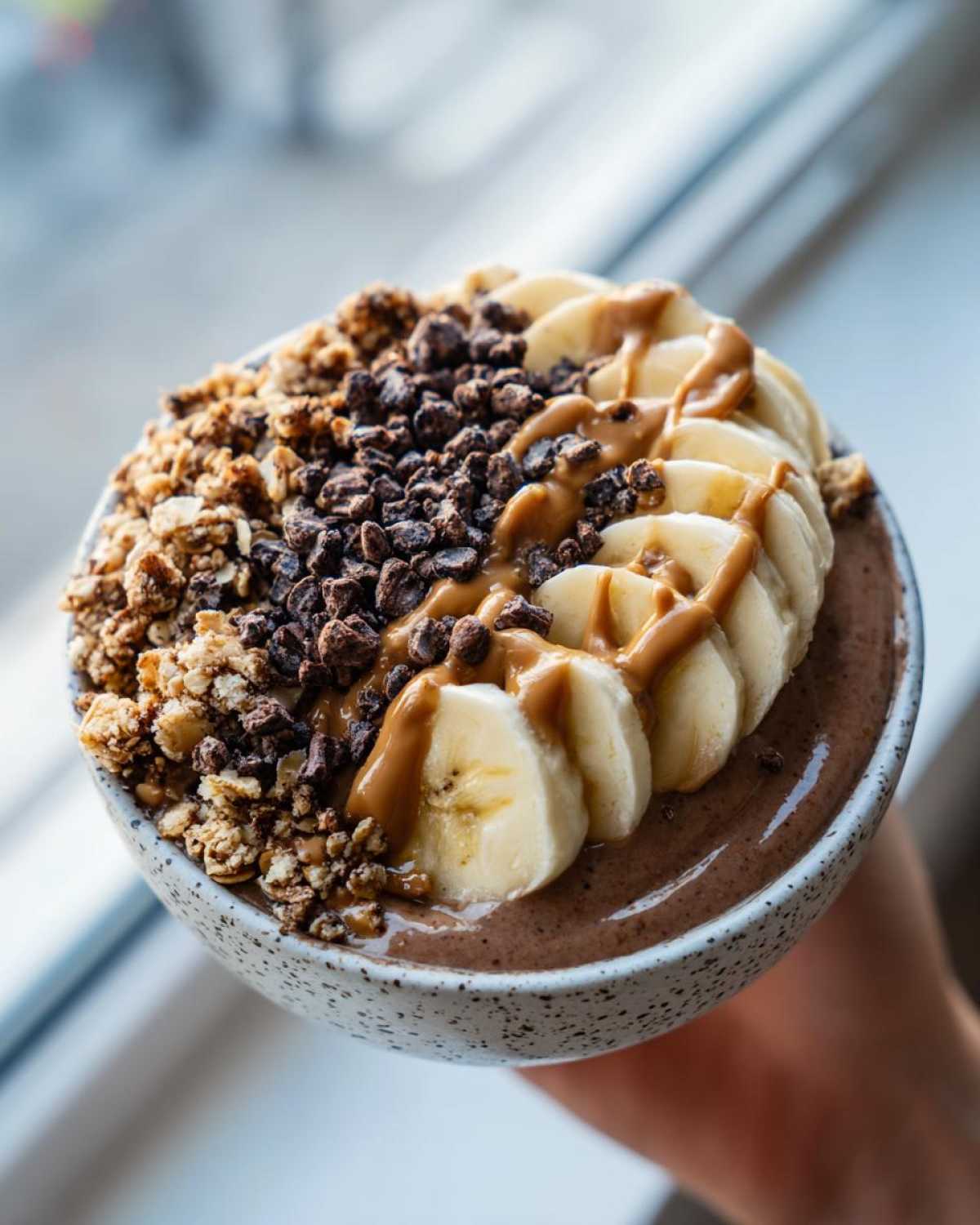 A close-up of a thick Vegan Chocolate Peanut Butter Banana Smoothie Bowl topped with sliced bananas, granola, and peanut butter drizzle.