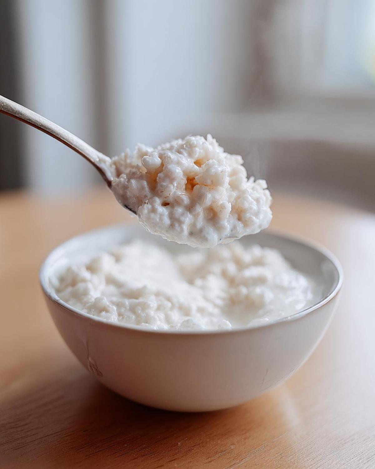 A spoonful of hot Vegan Creamy Coconut Rice Pudding being lifted from a white bowl on a wooden table.