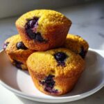 Three golden-brown Vegan Lemon Blueberry Muffins stacked on a white plate, bursting with dark blueberries.