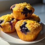 A stack of four golden Vegan Lemon Blueberry Muffins with visible blueberries and sugar topping on a white plate.