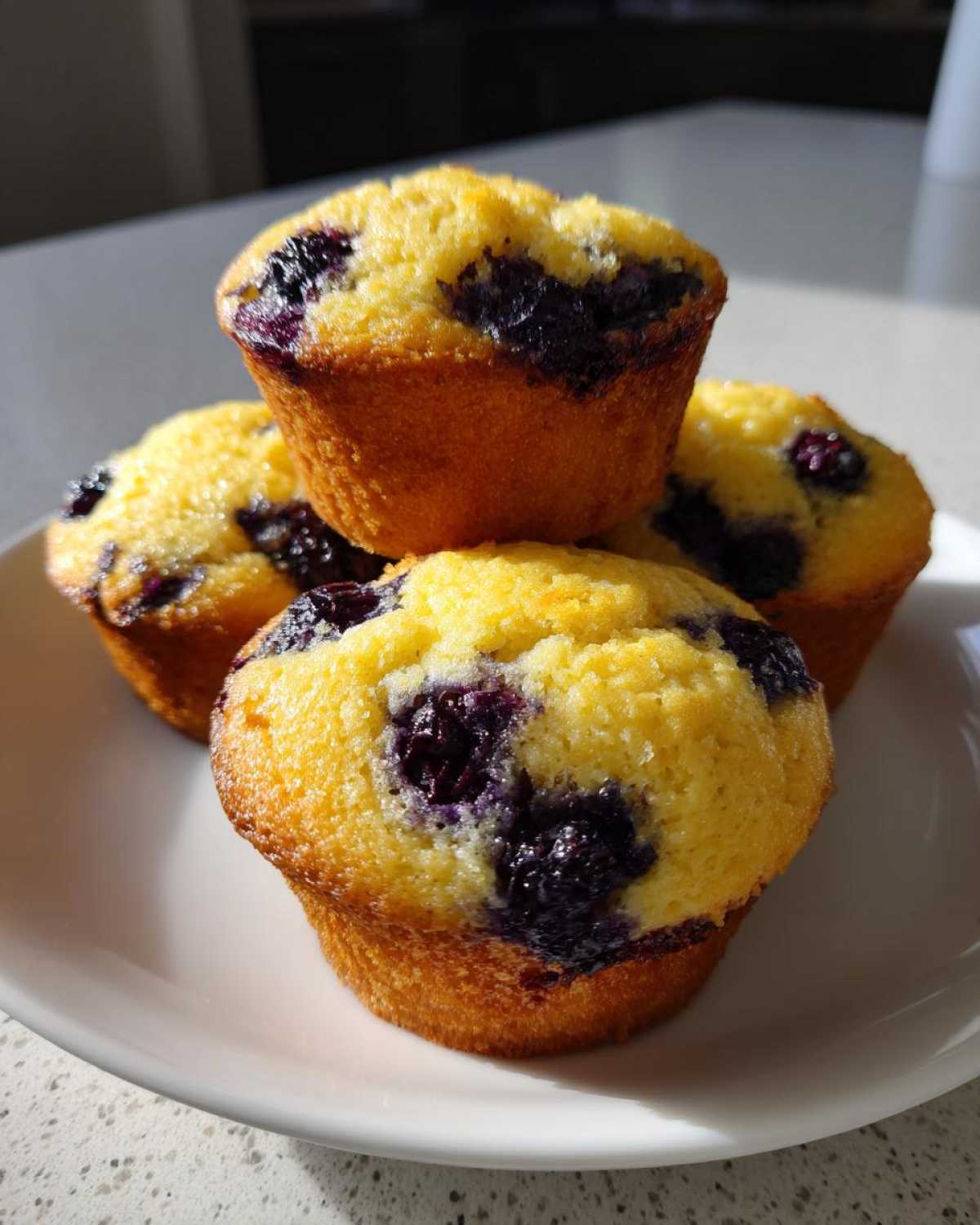 A stack of four golden Vegan Lemon Blueberry Muffins studded with dark blueberries, resting on a white plate.
