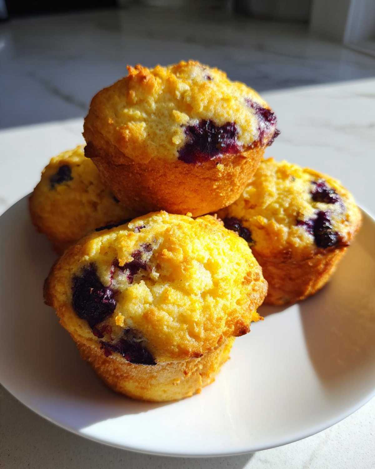 Four golden-brown Vegan Lemon Blueberry Muffins piled on a white plate, showing bright lemon color and dark blueberry bursts.
