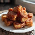 A pile of golden Air Fryer Crispy Cinnamon French Toast Bites coated heavily in cinnamon sugar on a white plate.