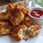 A serving of golden brown Air Fryer Crispy Coconut Chicken Bites on a white plate with a side of red dipping sauce.