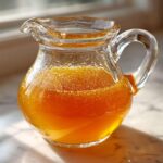 A small glass pitcher filled with bright orange Copycat Apple Cider Vinaigrette, sitting on a marble surface near a window.