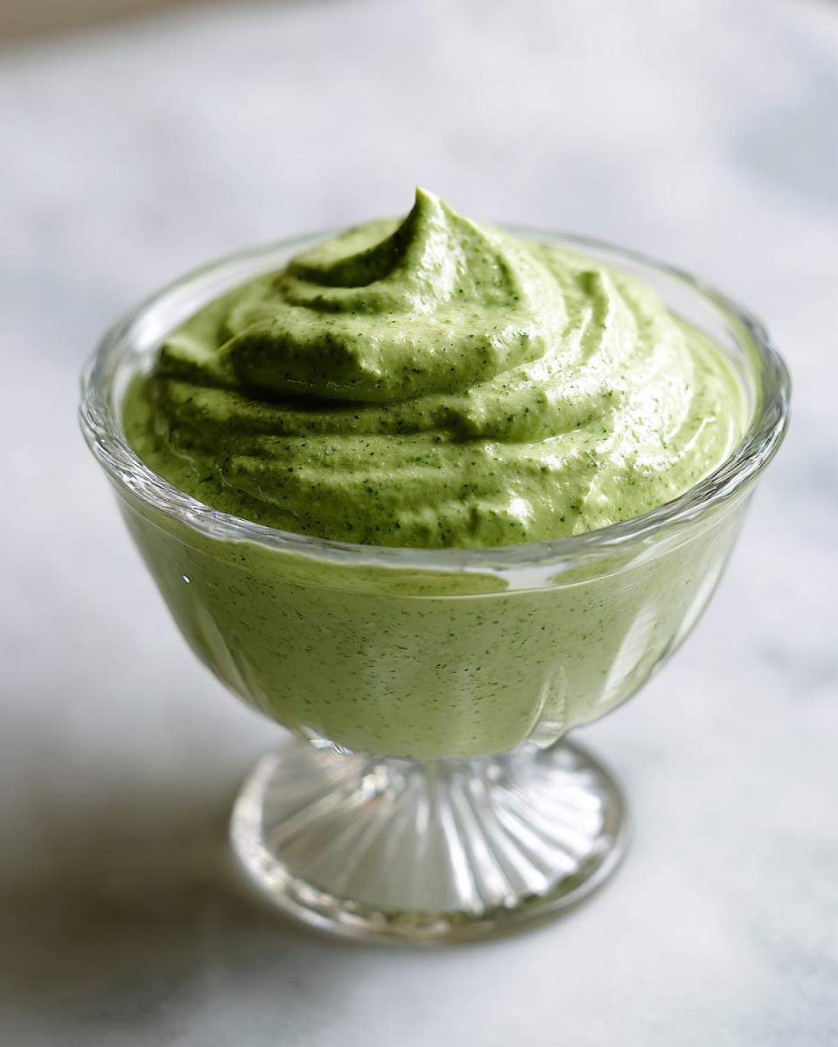 A close-up of thick, vibrant green Copycat Green Goddess Ranch Dressing swirled high in a small, clear glass serving dish.