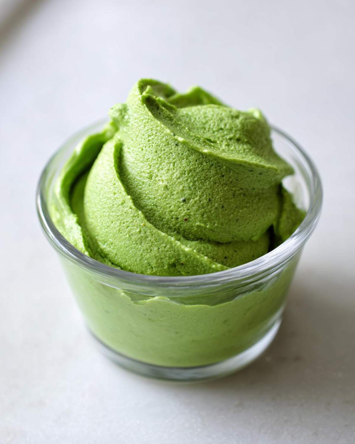 A close-up of thick, vibrant green Copycat Green Goddess Ranch Dressing swirled high in a small clear glass jar.