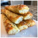 A stack of golden, soft Copycat Italian Restaurant Soft Garlic Breadsticks sprinkled with herbs and salt.