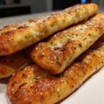 A stack of golden brown Copycat Italian Restaurant Soft Garlic Breadsticks topped with herbs and Parmesan cheese.
