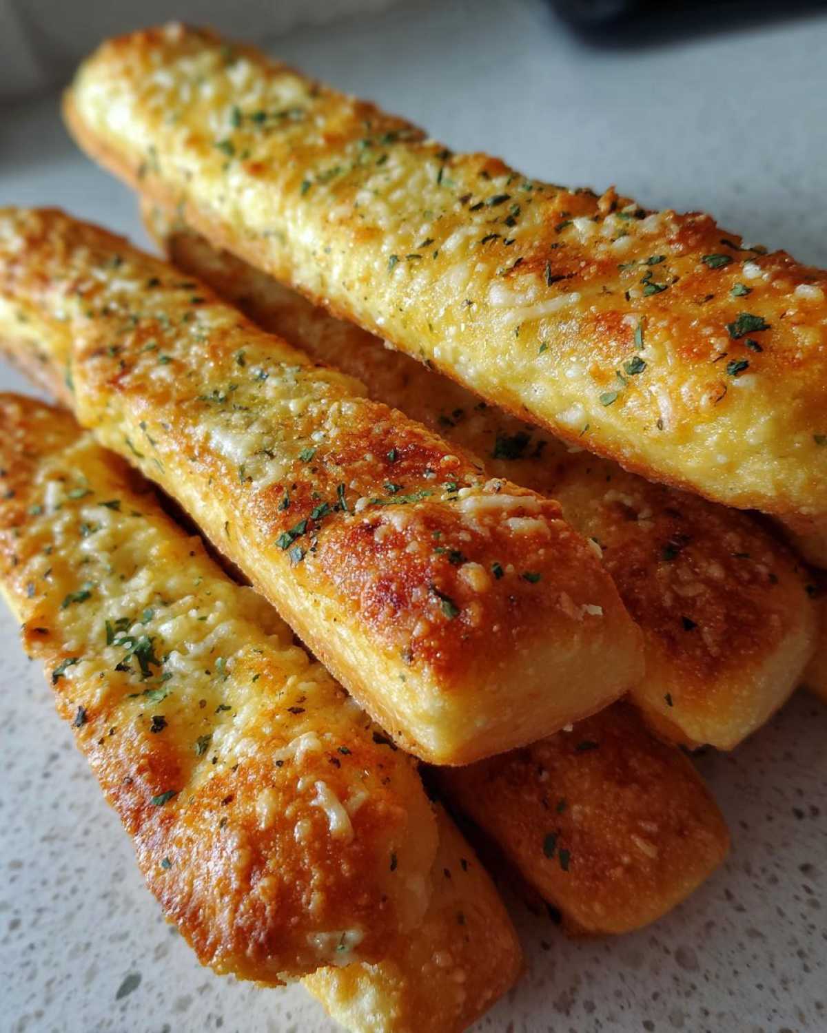 Close-up of several golden Copycat Italian Restaurant Soft Garlic Breadsticks stacked together, topped with melted cheese and parsley.