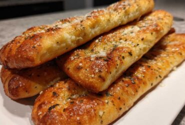 A stack of golden brown Copycat Italian Restaurant Soft Garlic Breadsticks topped with herbs and Parmesan cheese.
