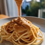 Close-up of thick, creamy Easy 5-Minute Peanut Sauce being poured over a mound of spaghetti noodles on a white plate.