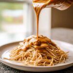 A thick stream of Easy 5-Minute Peanut Sauce for Noodles being poured over a mound of cooked noodles on a white plate.