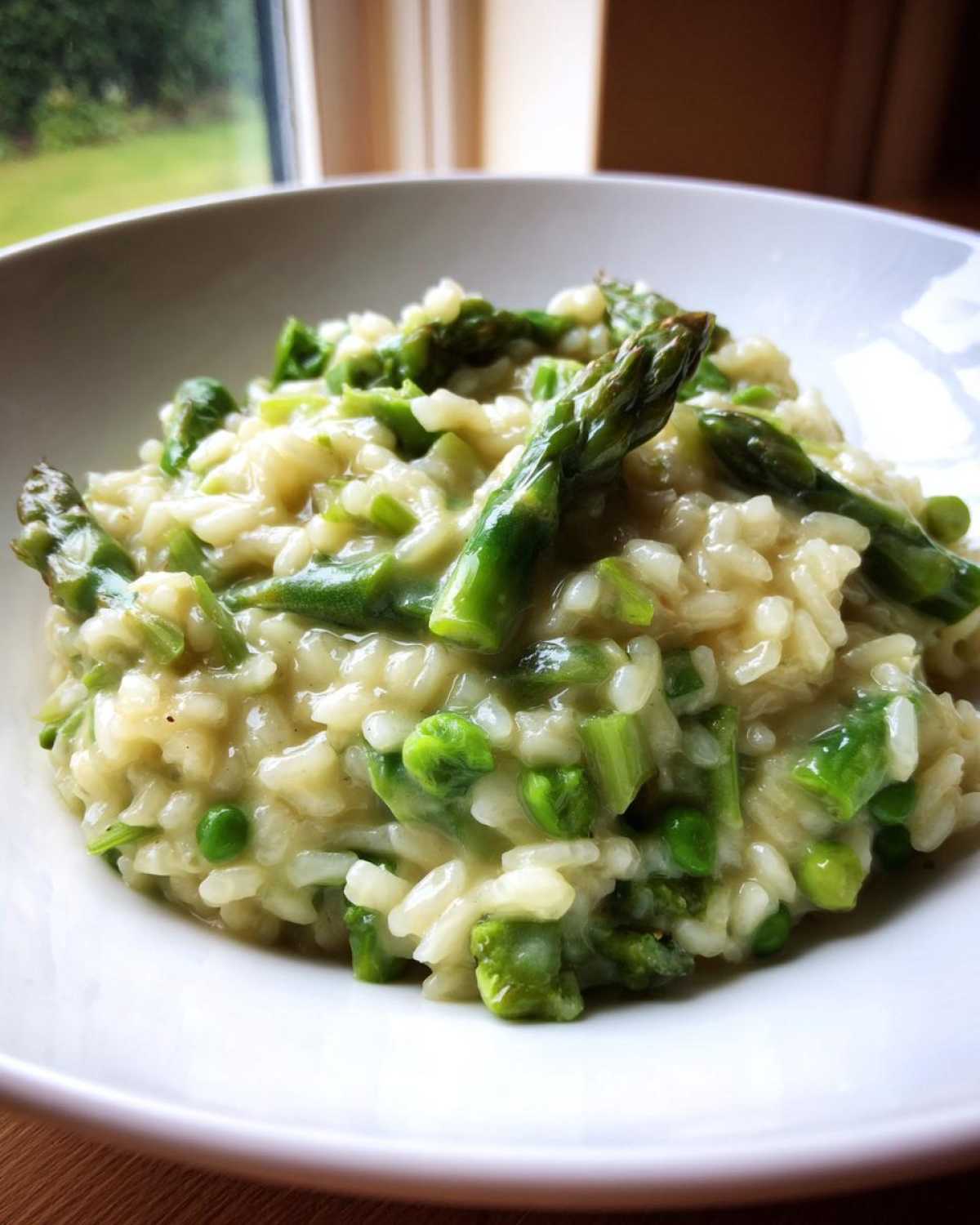 A close-up of a white bowl filled with Easy Creamy Asparagus Risotto, featuring bright green asparagus tips and peas.