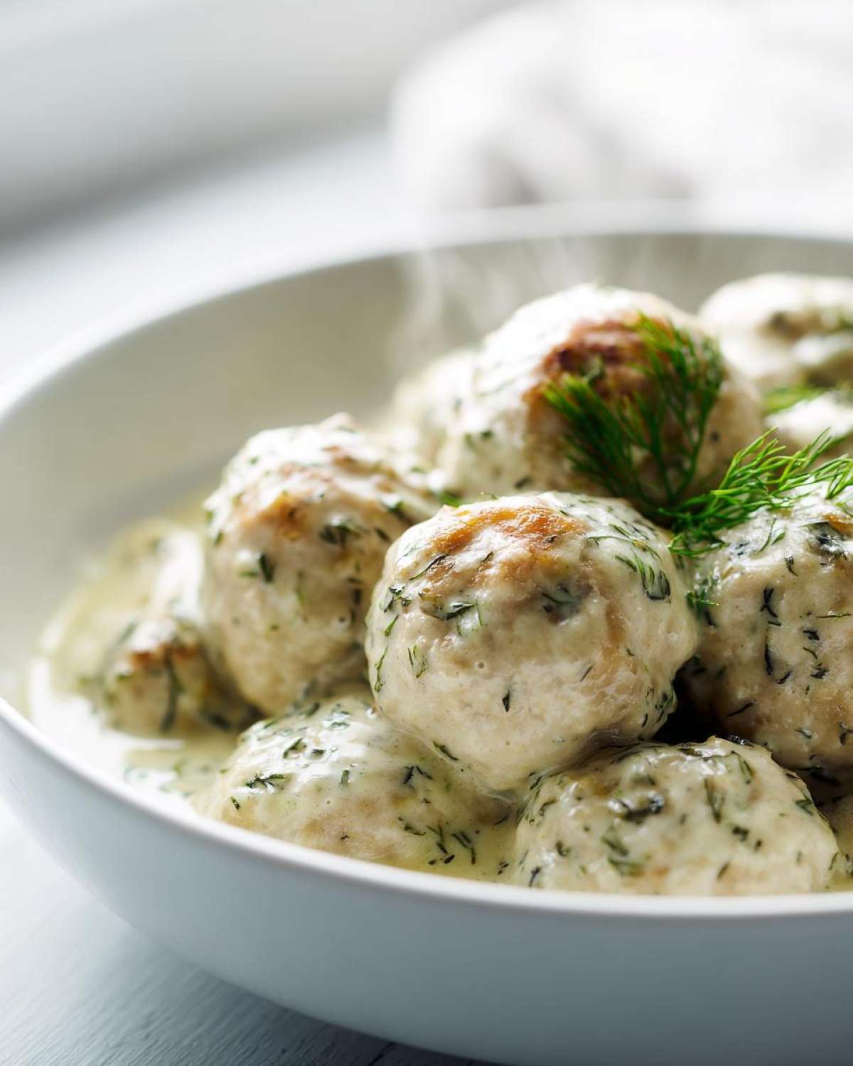 Close-up of hot Easy Creamy Greek Chicken Meatballs covered in a light dill sauce in a white bowl.