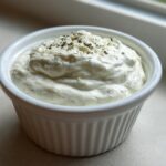 A small white ramekin filled with thick, creamy Easy Greek Yogurt Caesar Dressing, topped with grated cheese and herbs.