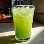 A tall glass filled with bright green, bubbly Easy Iced Cucumber Lime Agua Fresca and ice cubes, lit by sunlight.