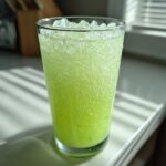 A tall glass filled with bright green Easy Iced Cucumber Lime Agua Fresca, topped with crushed ice.