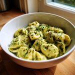 A close-up of a white bowl filled with Easy One-Pot Creamy Pesto Tortellini, topped with grated cheese and herbs.