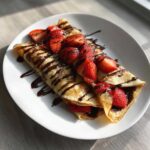 Two rolled Gluten-Free Chocolate Strawberry Crepes topped with fresh sliced strawberries and drizzled with chocolate sauce.