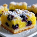 A close-up of a square Gluten-Free Lemon Blueberry Layer Bar, featuring a bright yellow lemon filling, whole blueberries, and a dusting of powdered sugar.