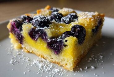 Close-up of a single serving of Gluten-Free Lemon Blueberry Layer Bars with bright yellow filling and dark blueberries, dusted with powdered sugar.